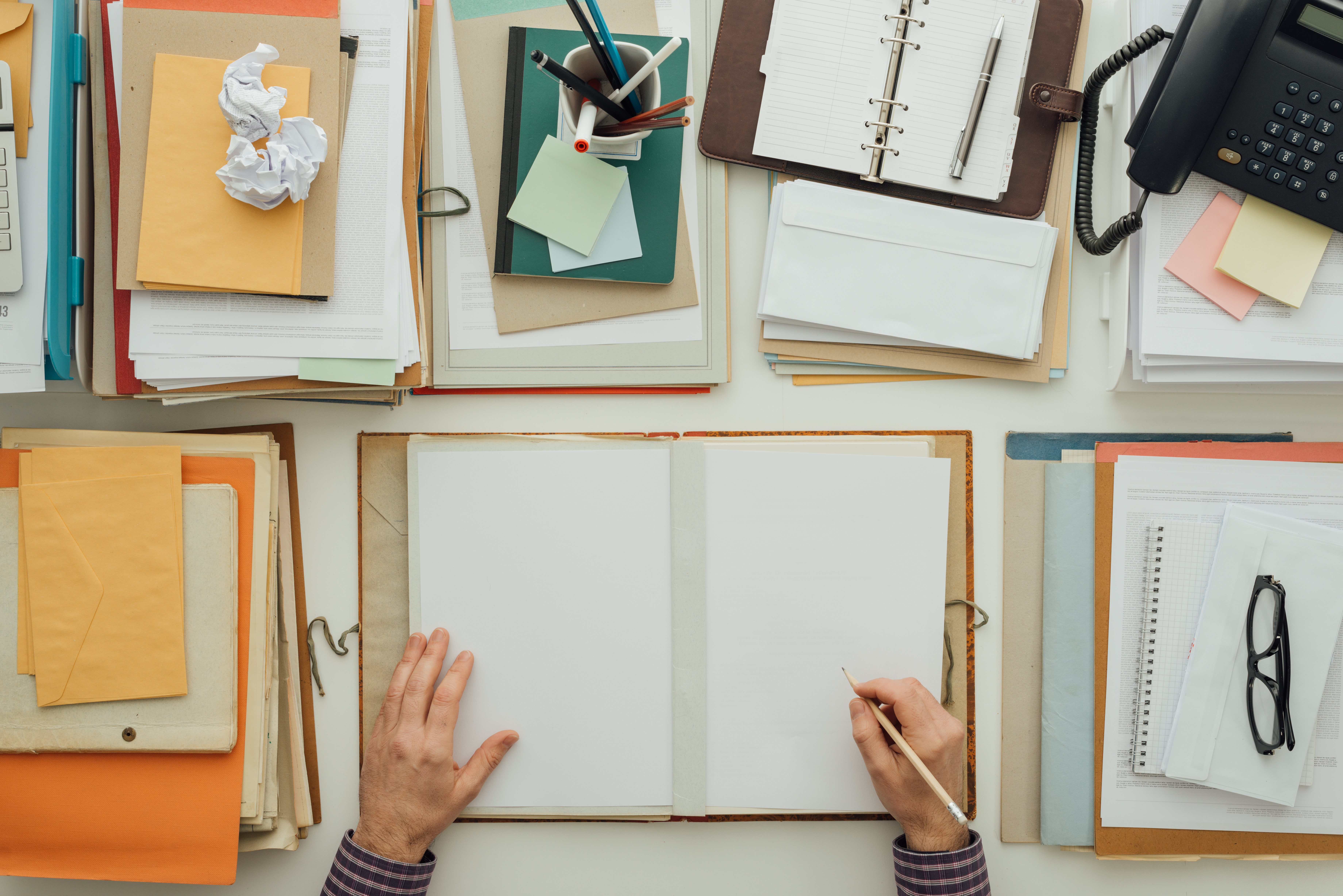 Office worker writing paperwork and documents in a folder on a full business desk,