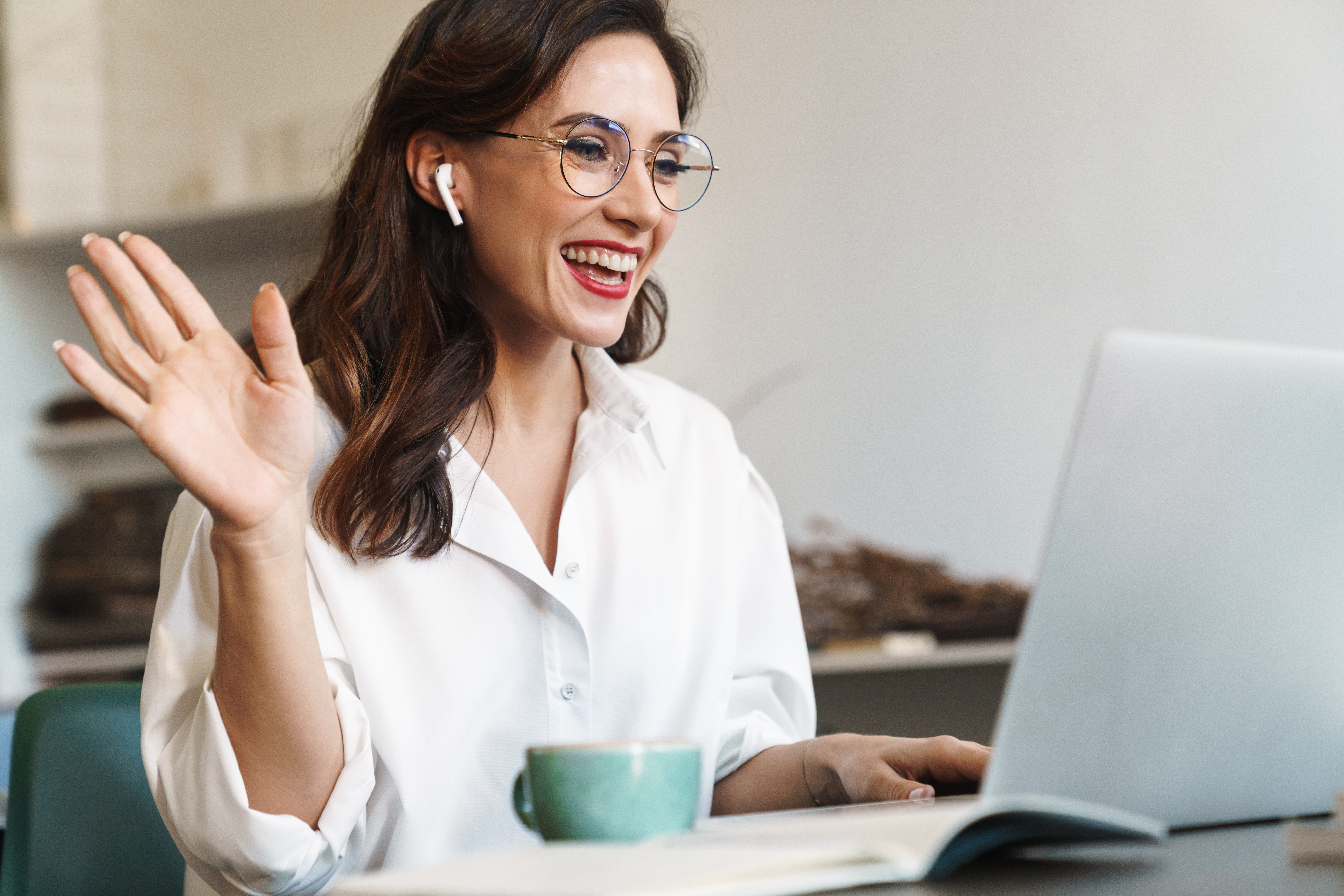 cheerful young brunette businesswoman sitting at the cafe table with laptop computer indoors, wearing wireless earphones, audio call, waving hand