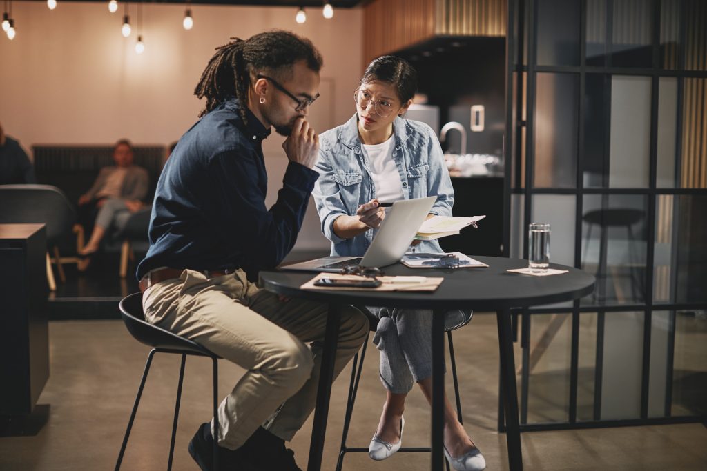 Two young businesspeople talking together over a laptop during a meeting around an office table